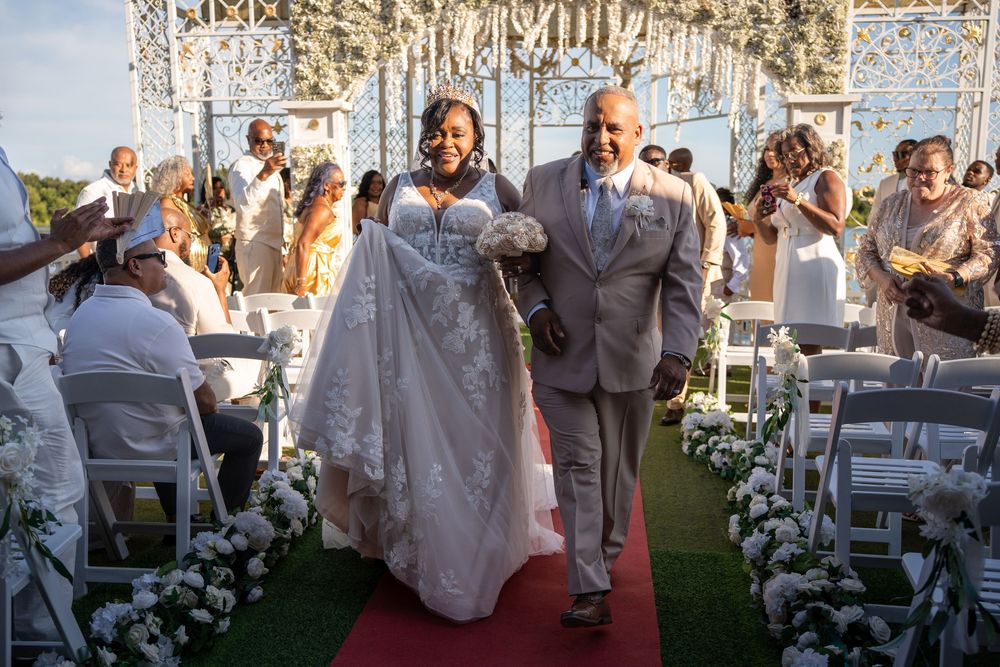 Bride in lace gown walks down red carpet aisle with escort at outdoor wedding ceremony.