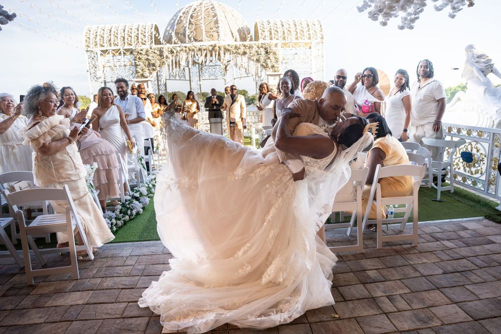Groom lifts bride in stunning white lace gown during outdoor wedding ceremony with elegant white gazebo backdrop.