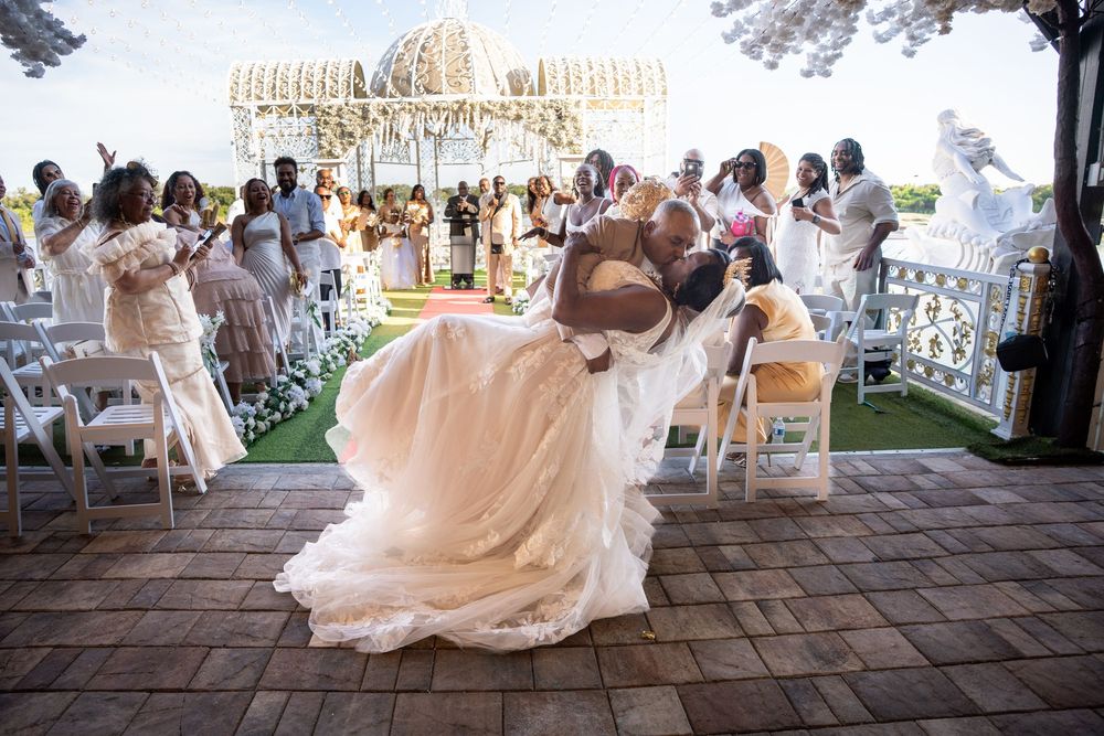 Bride and groom share a romantic dip kiss at outdoor wedding ceremony with guests cheering on red carpet aisle.