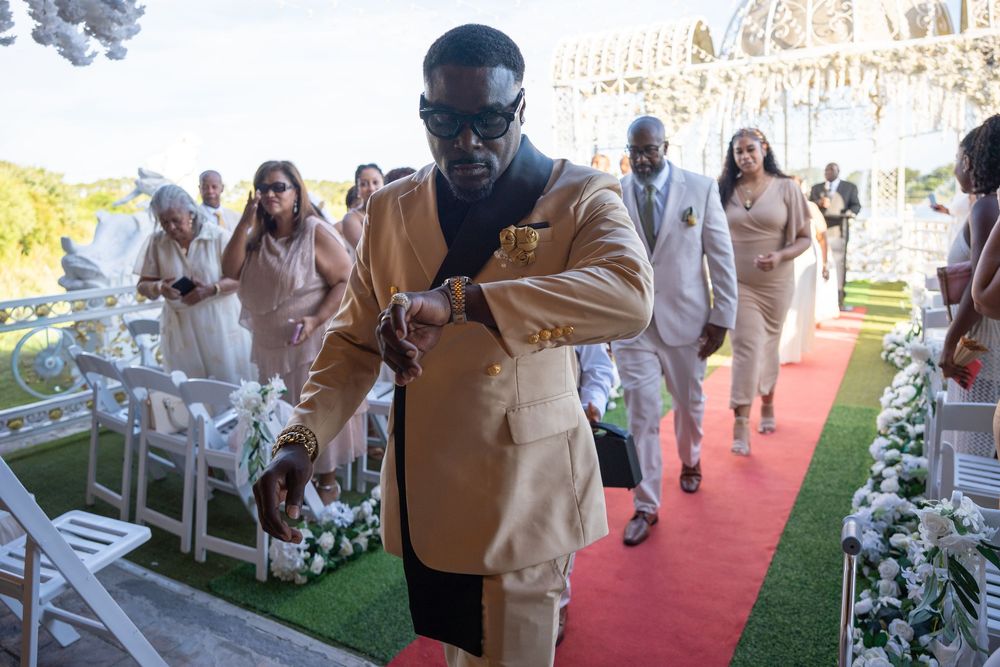 Elegantly dressed man in gold suit adjusts watch at outdoor wedding ceremony with guests and floral decor.