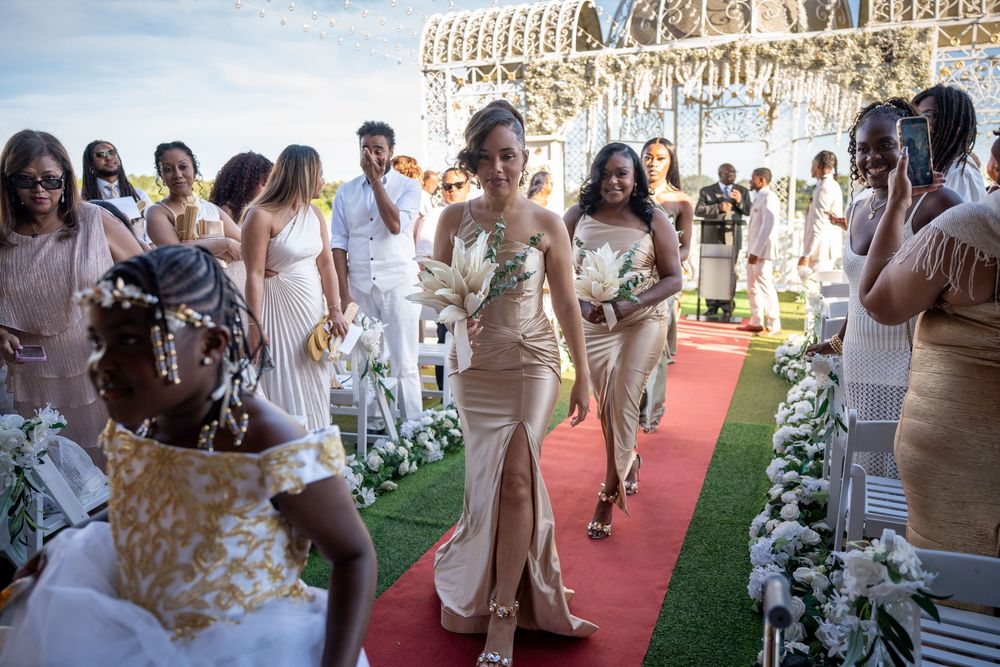 Bridesmaids in gold satin gowns walk down a red carpet aisle at an elegant outdoor wedding ceremony.