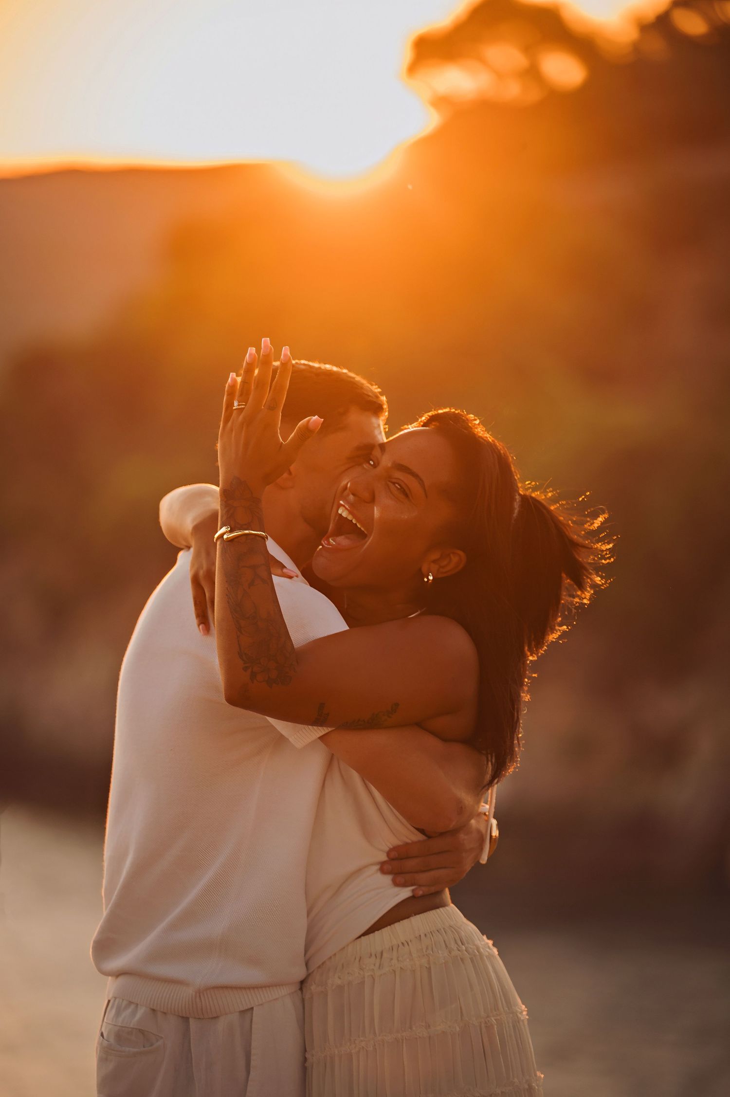 A joyful couple embraces at golden sunset, woman laughing as man kisses her cheek outdoors.