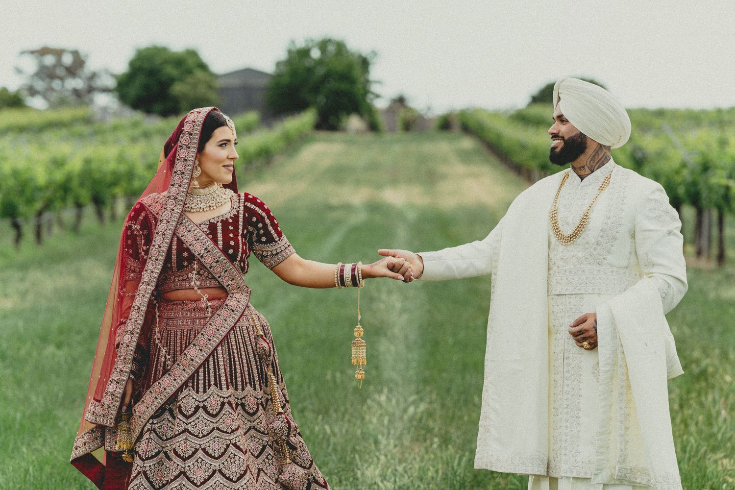 Indian couple in traditional wedding attire holding hands in a vineyard, bride in red lehenga, groom in white sherwani.