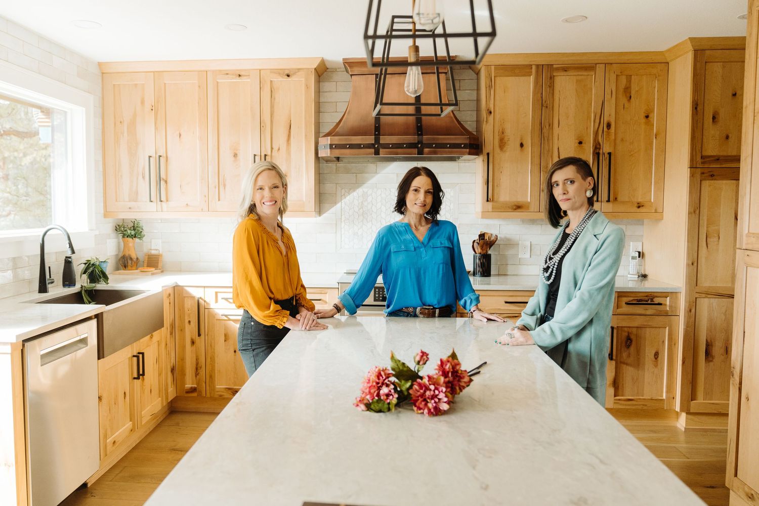 Three women smile around a marble kitchen island with wood cabinets and a statement range hood.