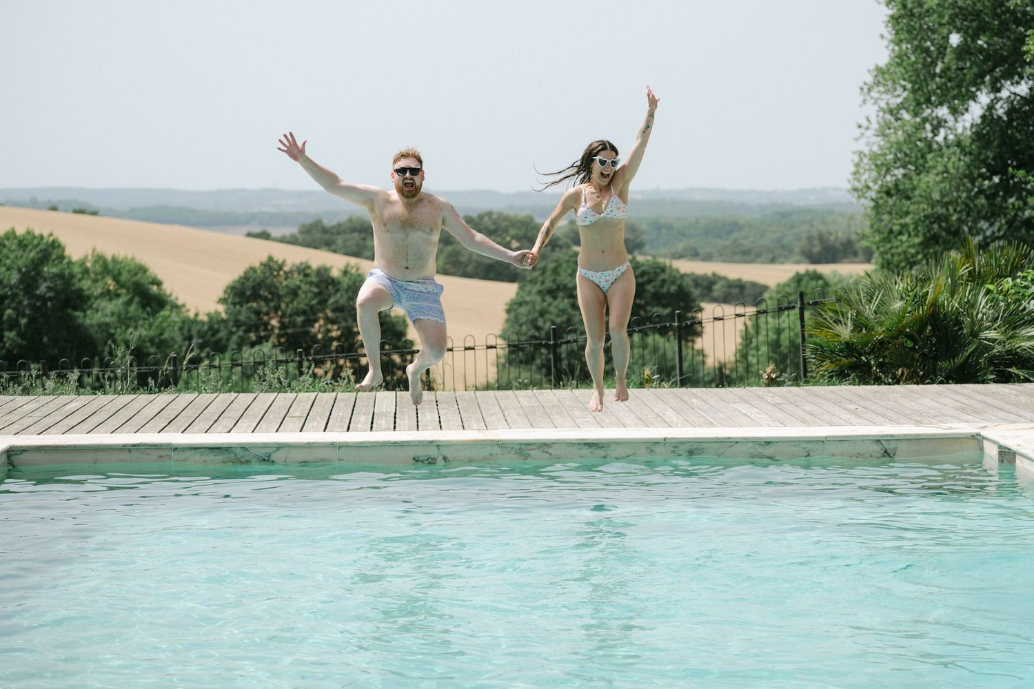 Couple jumping into outdoor pool holding hands, surrounded by lush greenery and scenic countryside views.