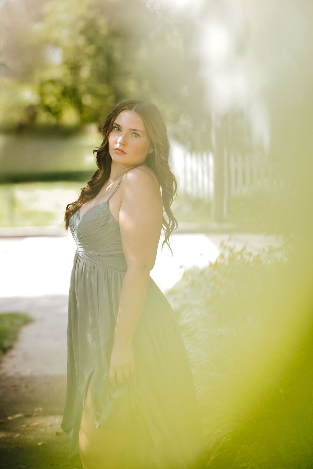 Young woman in a sage green sundress poses gracefully outdoors in a sunlit garden setting.