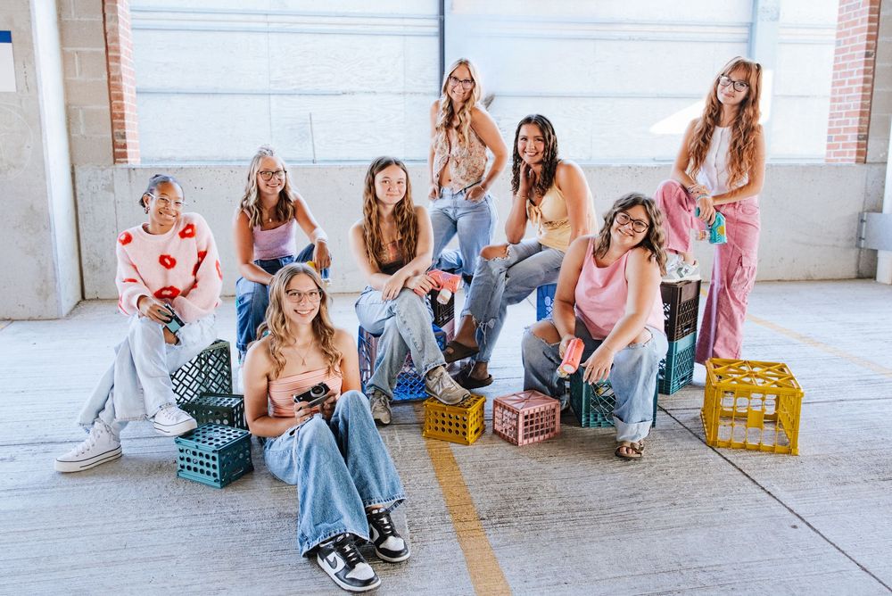 Group of nine young women posing together on colorful milk crates in a bright industrial space.