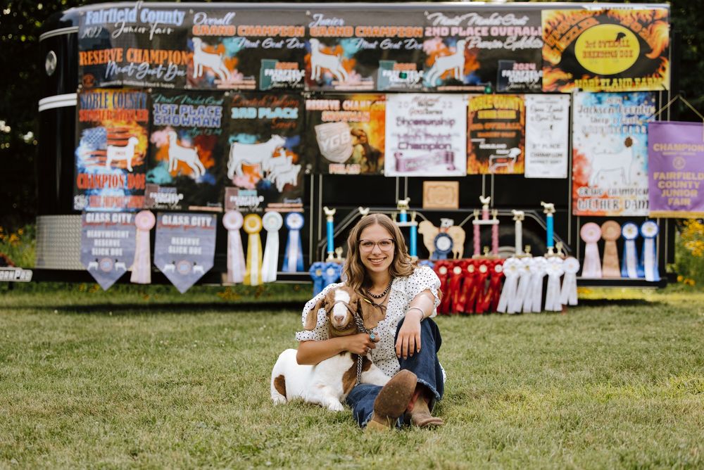 Young girl poses with her goat and award ribbons at a county fair livestock competition on green grass.