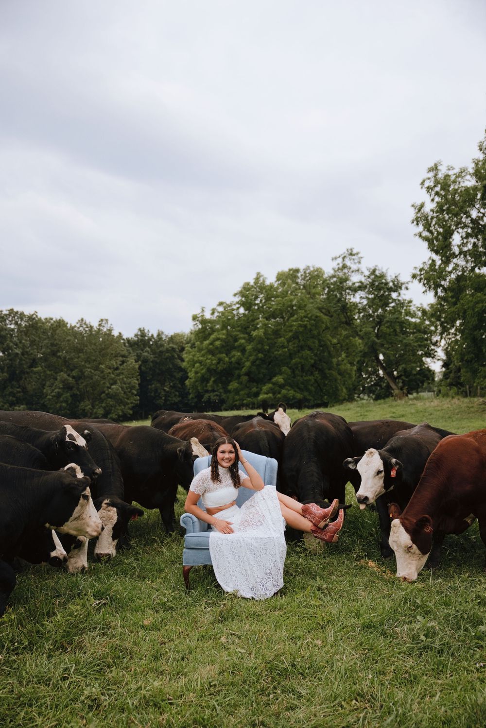 A smiling woman in a white dress sits in a blue chair surrounded by Hereford cattle in a lush green pasture.