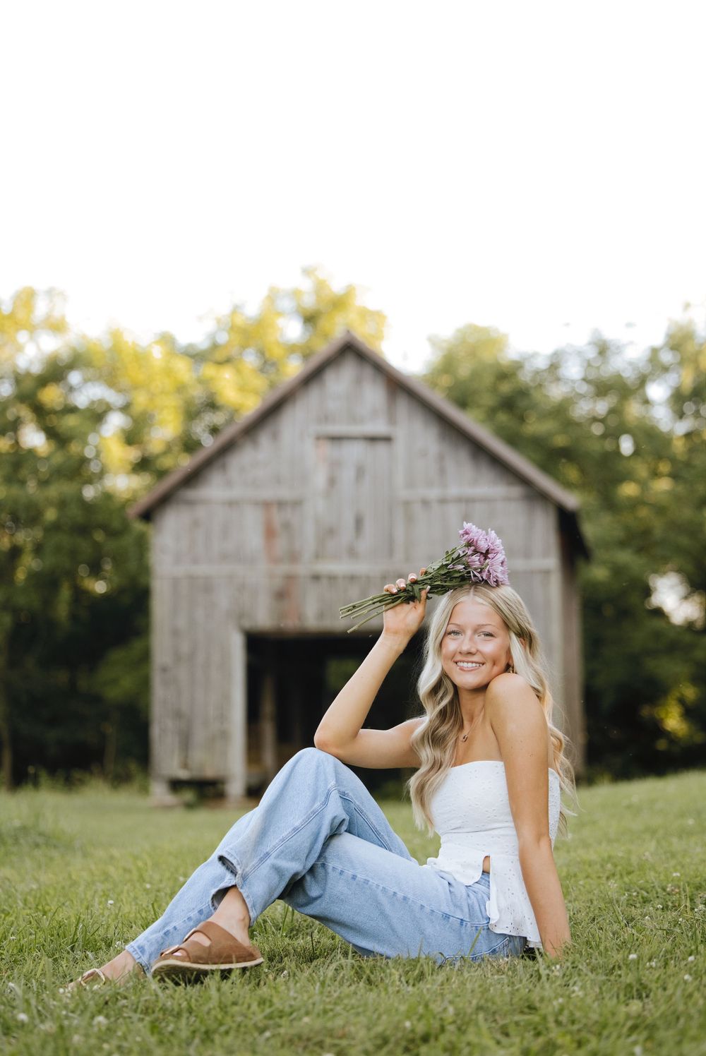 Young woman sitting on grass holding purple wildflowers, smiling in front of a rustic barn, wearing jeans and white top.
