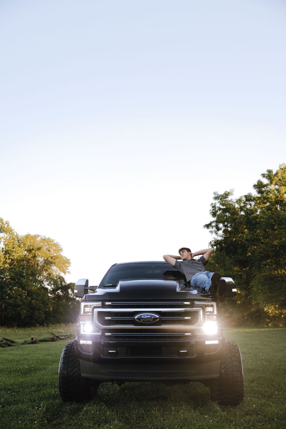 Person relaxing on hood of black Ford truck with headlights on, parked on green grass at dusk.