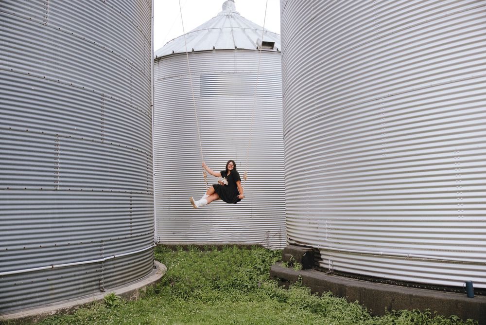 Woman sitting on a swing between large metal grain silos on a farm.