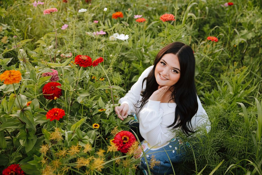 Young woman with dark hair sits smiling among vibrant red and orange zinnia flowers in a lush green garden.