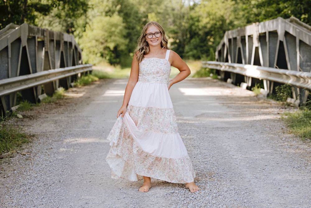 Young girl in white floral maxi dress and glasses poses barefoot on rustic wooden bridge surrounded by lush greenery.