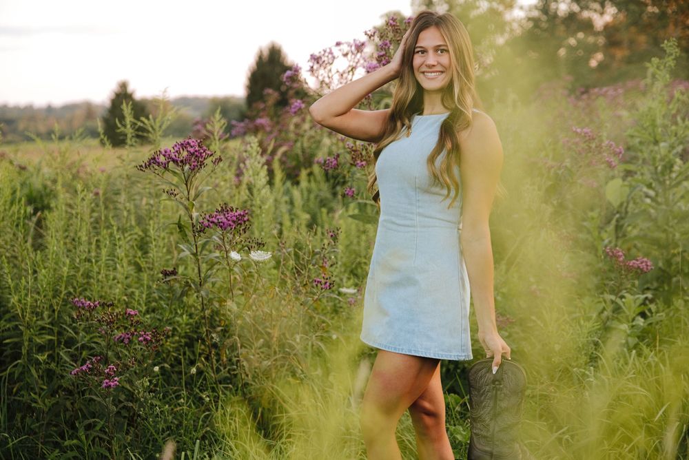 Young woman in light blue dress smiling among wildflowers in a lush green field on a sunny day.