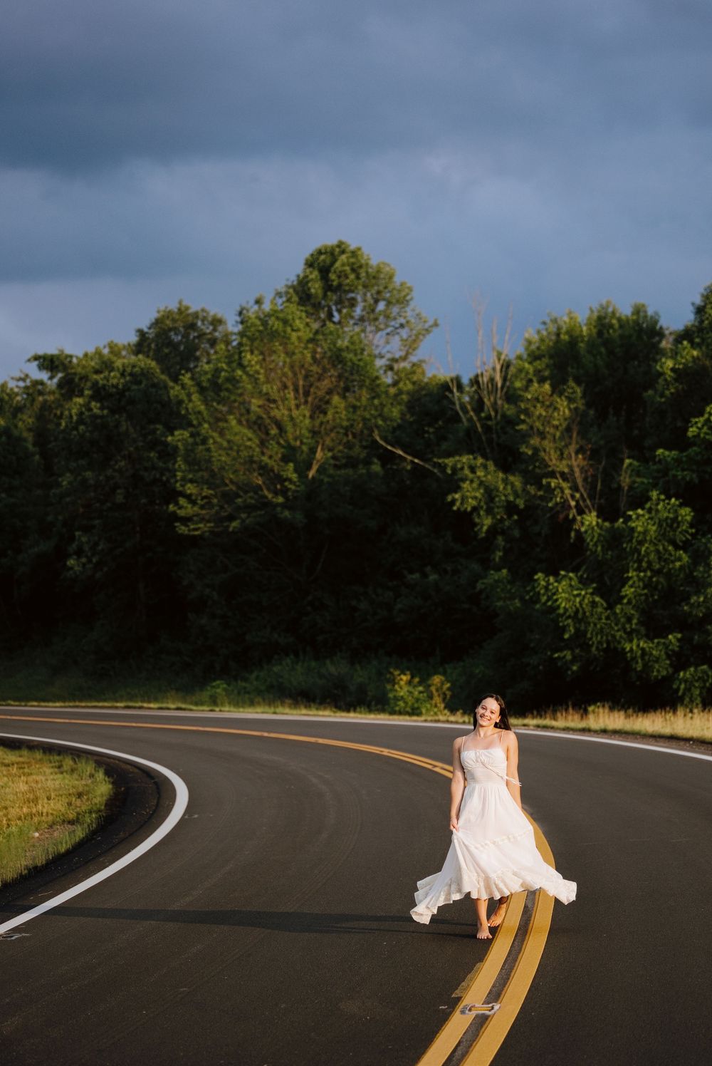 Woman in white dress walking on curved road surrounded by lush green trees under dramatic cloudy sky.