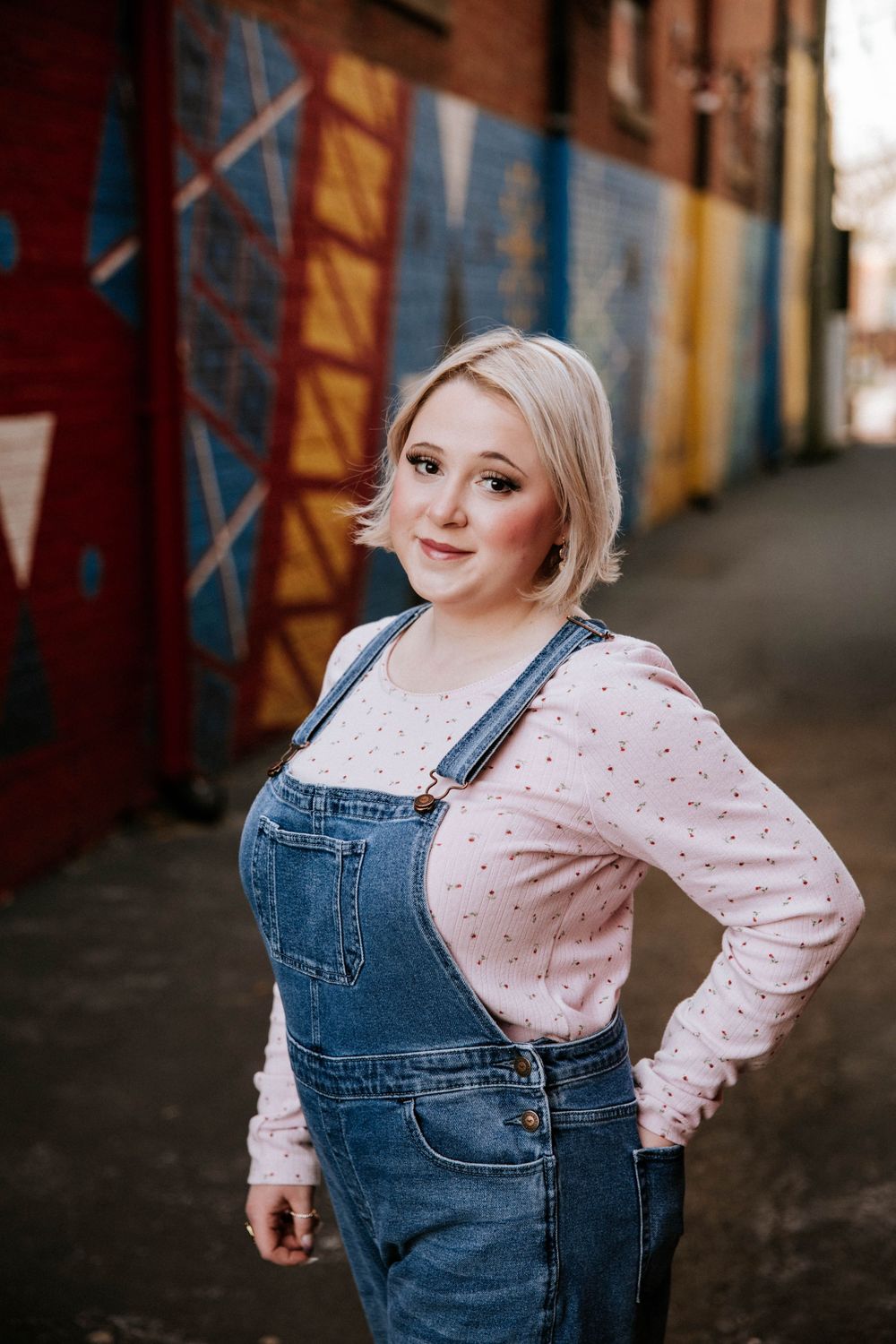Young blonde woman in denim overalls and pink shirt poses in colorful mural alley.