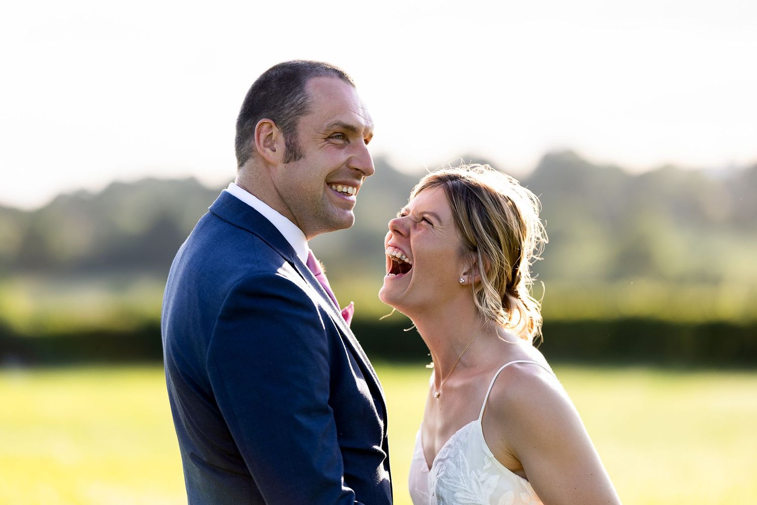 Laughing bride and groom in wedding attire share a joyful moment outdoors with lush green fields in the background.