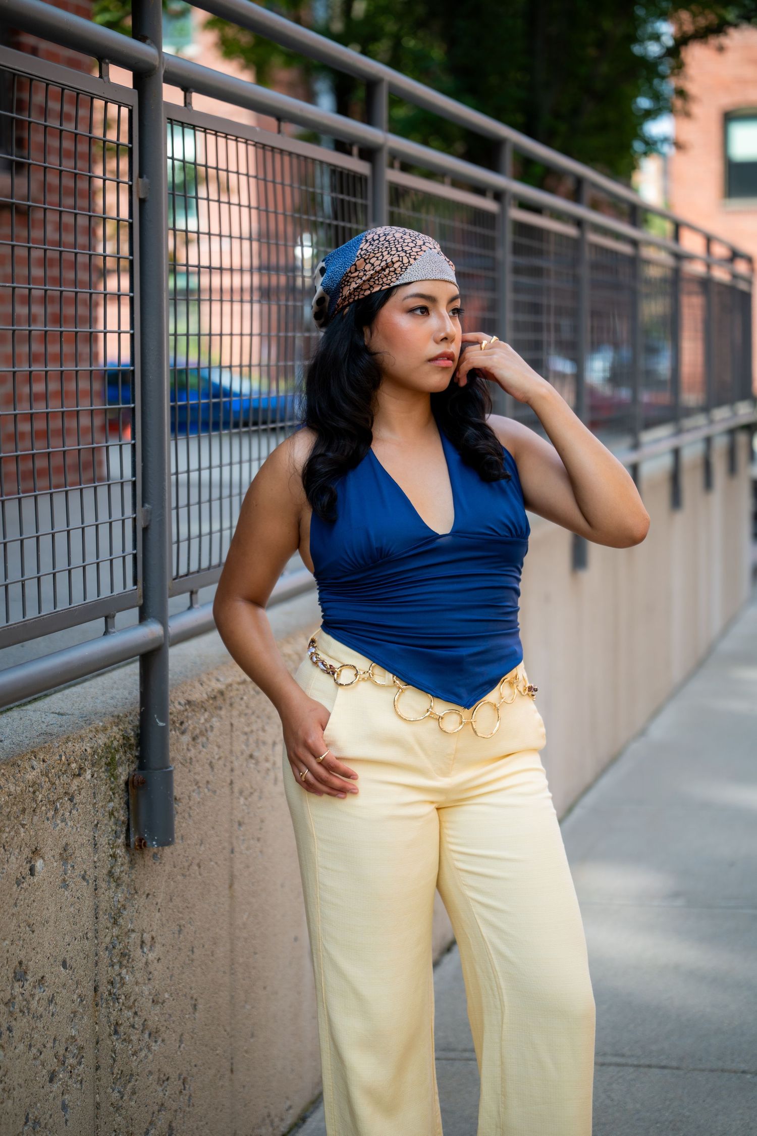 A young woman in a blue halter top, cream wide-leg pants, gold chain belt, and patterned headband poses on an urban sidewalk.