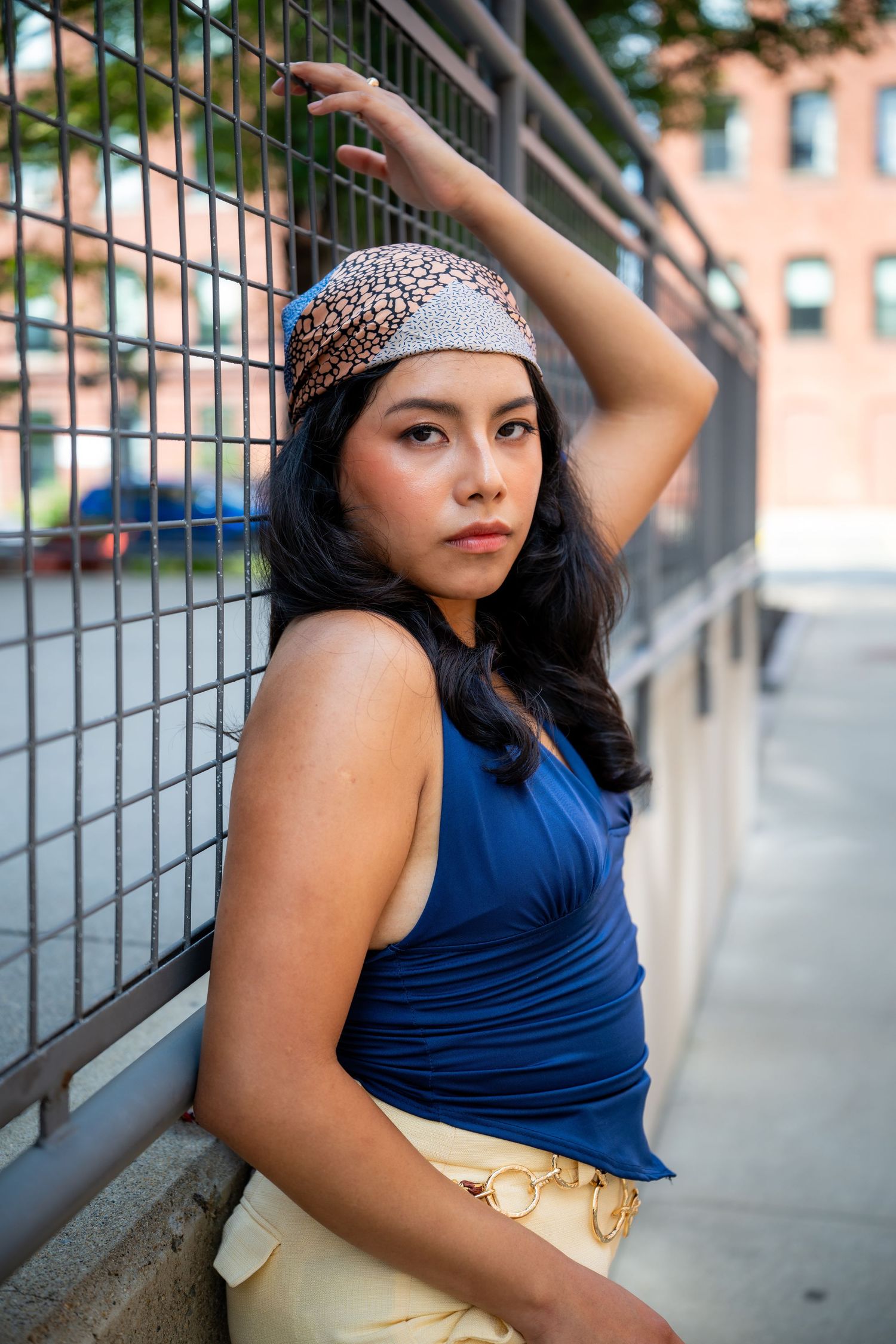 Young woman in blue top and headband poses against urban metal fence with brick building backdrop.