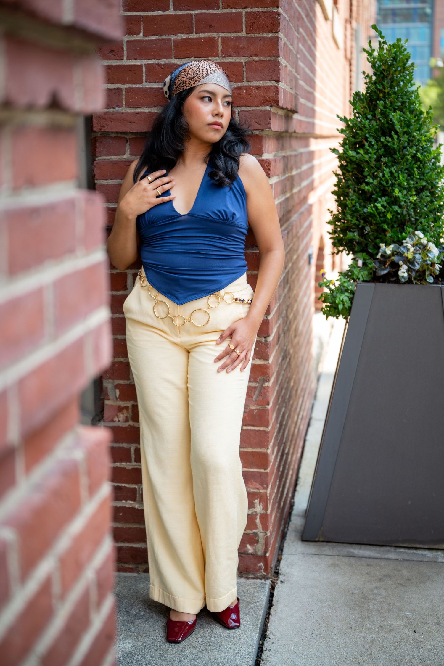 Woman in blue halter top, cream wide-leg pants, and red heels poses against a brick wall outdoors.