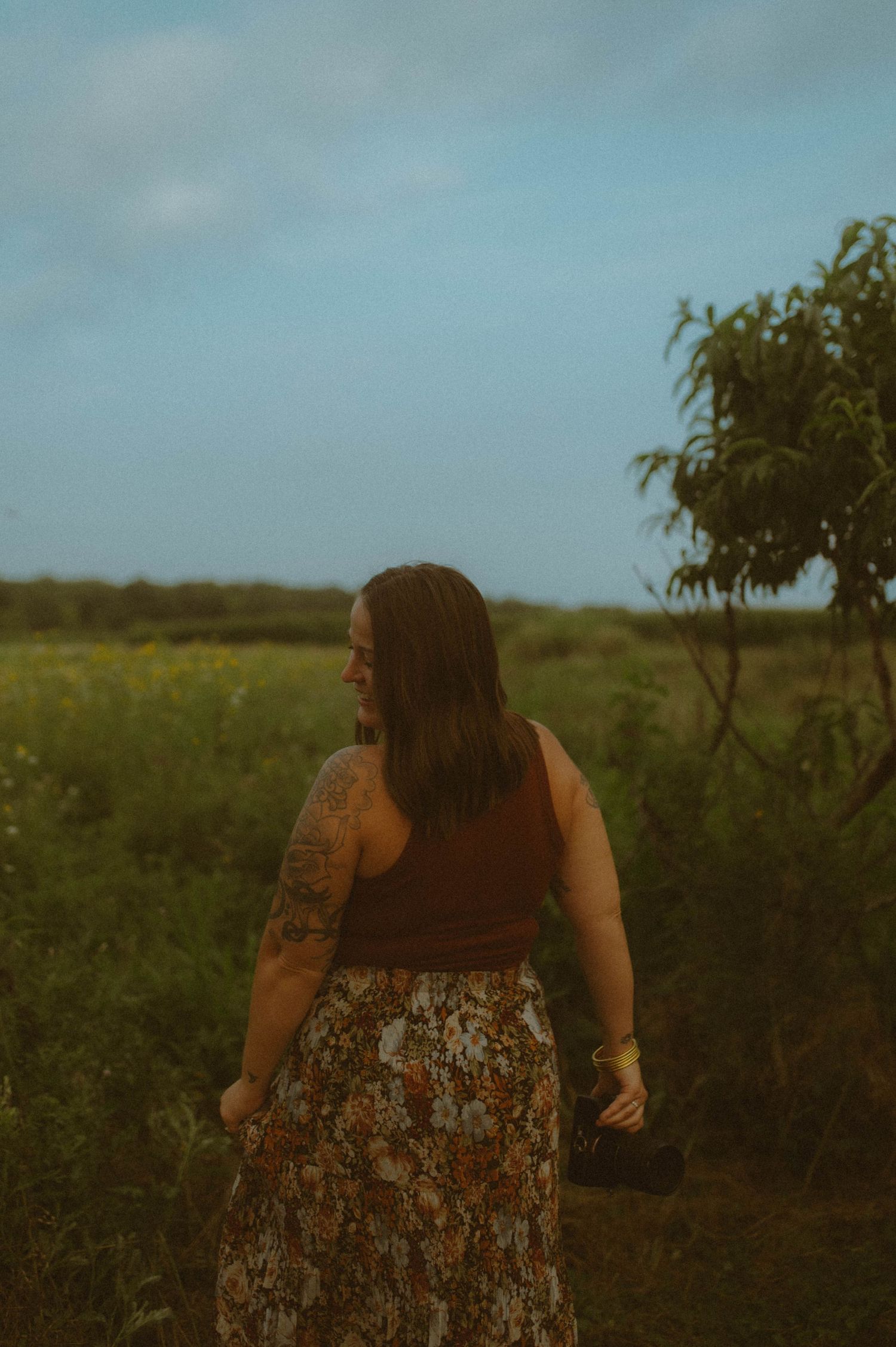 A tattooed woman in a brown top and floral skirt walks through a green field.