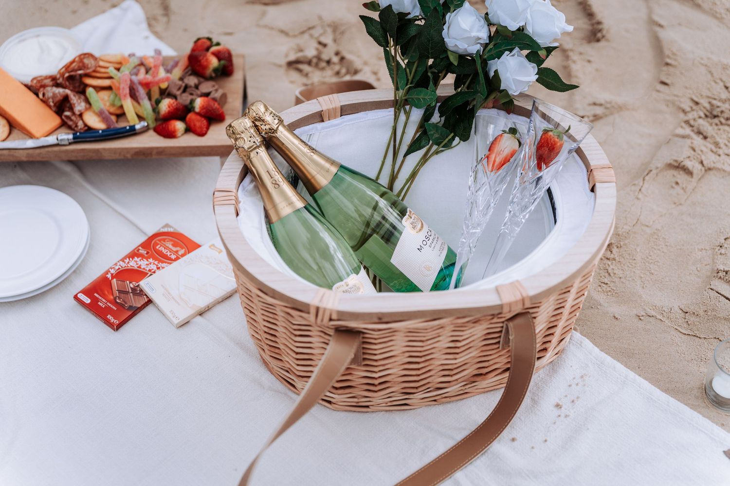 Wicker picnic basket with champagne bottles and white roses on sandy beach.