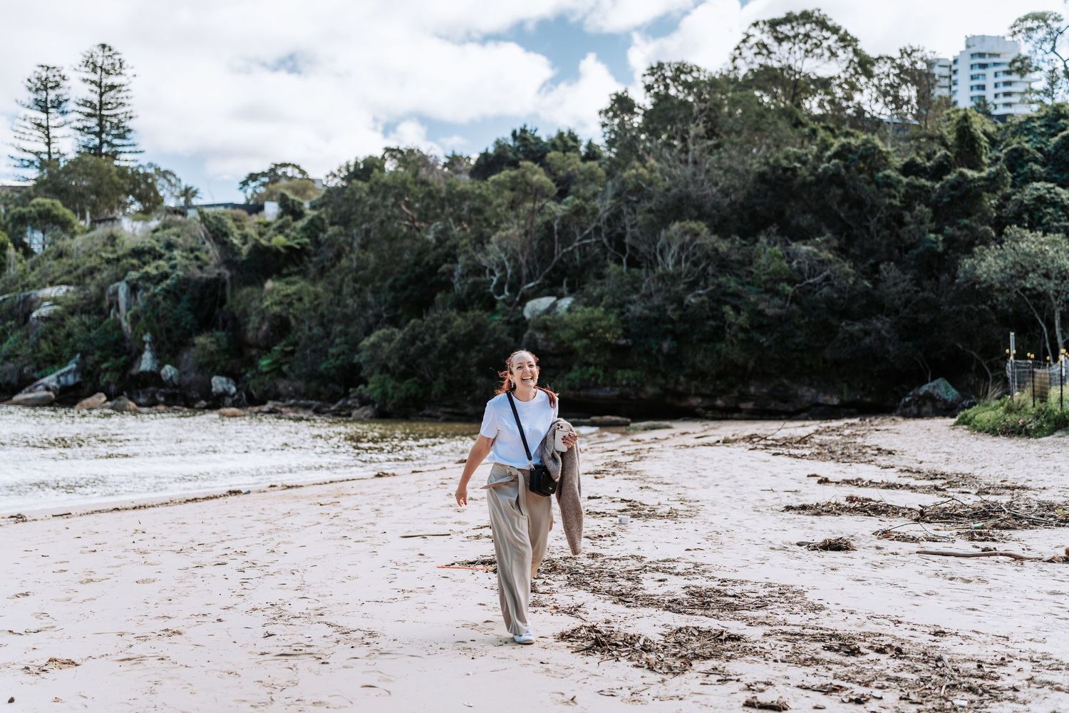 A man walks along a sandy beach with lush green trees in the background.