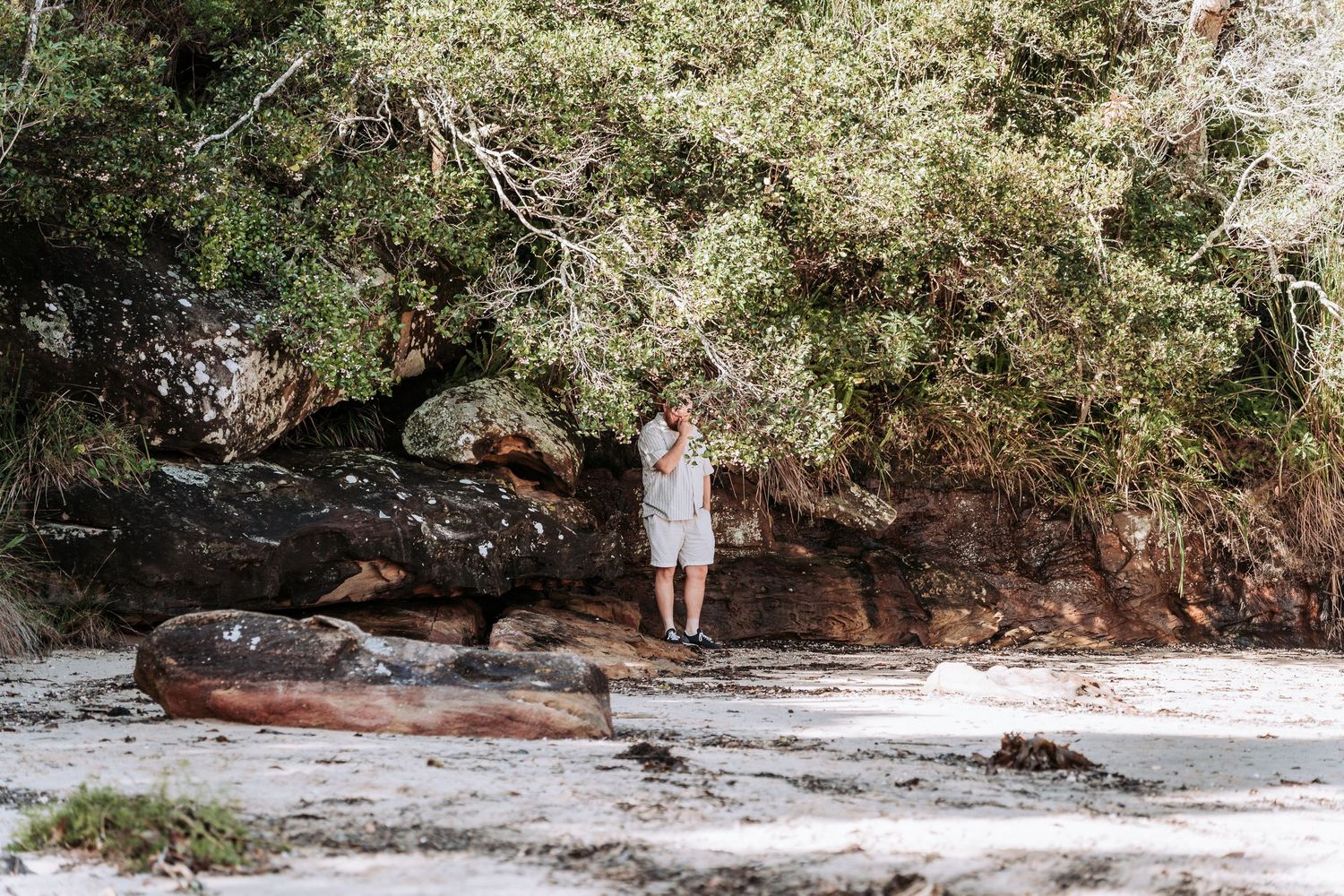 Person standing among large rocks and lush green trees on a sandy beach.