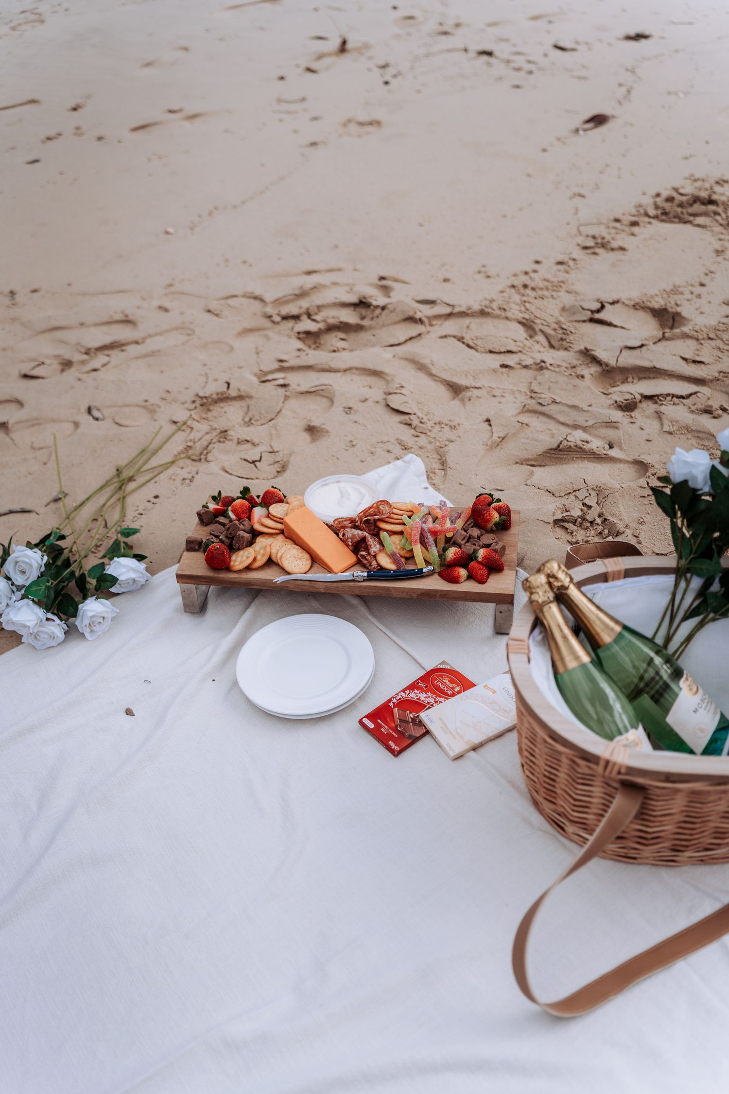 Beach picnic setup with charcuterie board, strawberries, champagne in wicker basket, and white blanket on sandy shore.