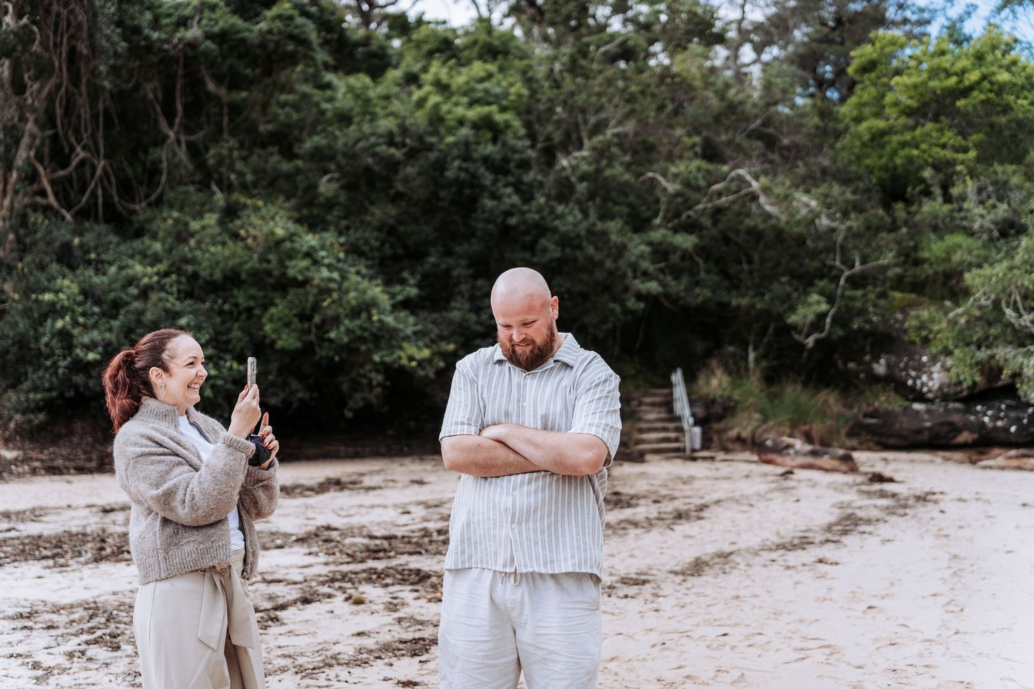 Woman photographing man with arms crossed on sandy beach with lush green trees behind.