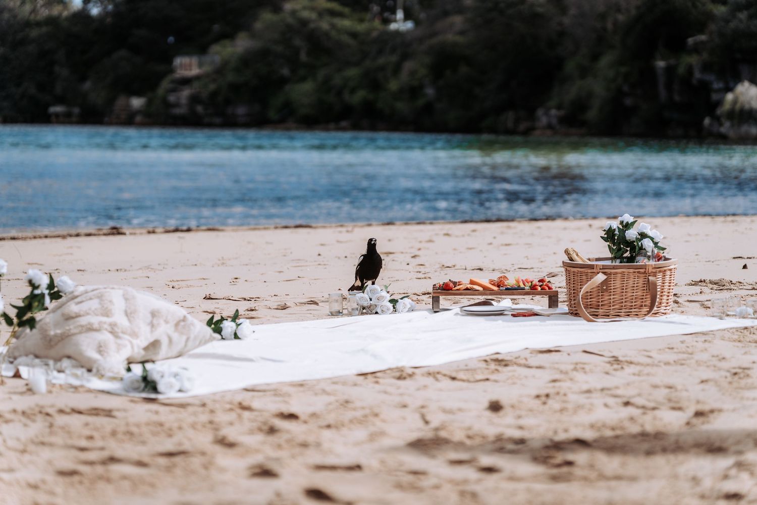 A romantic beach picnic setup with a wicker basket, white blanket, and flowers by the water.