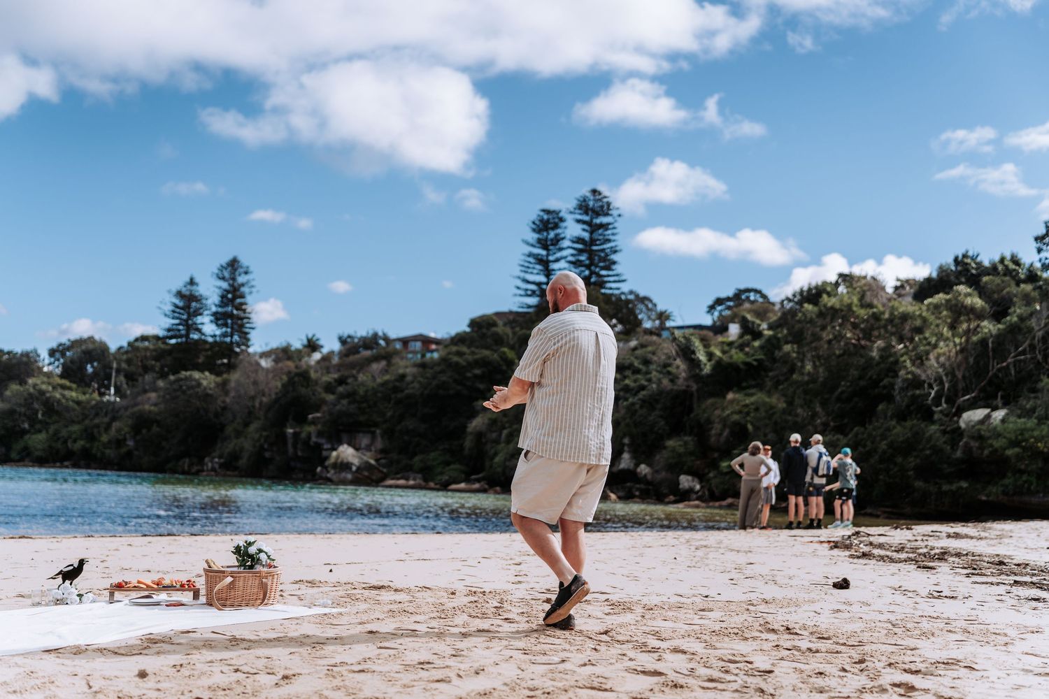 Bald man playing cricket on a sandy beach with trees and blue sky in background, Australia.