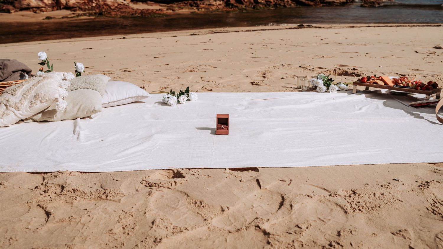 White picnic blanket with decorations set up on a sandy beach for a romantic outdoor celebration.