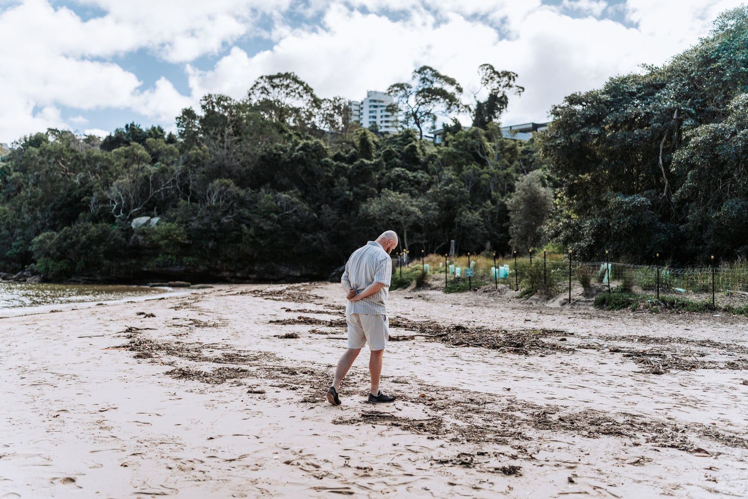 Elderly man walking on sandy beach with lush green trees and cloudy sky in background.