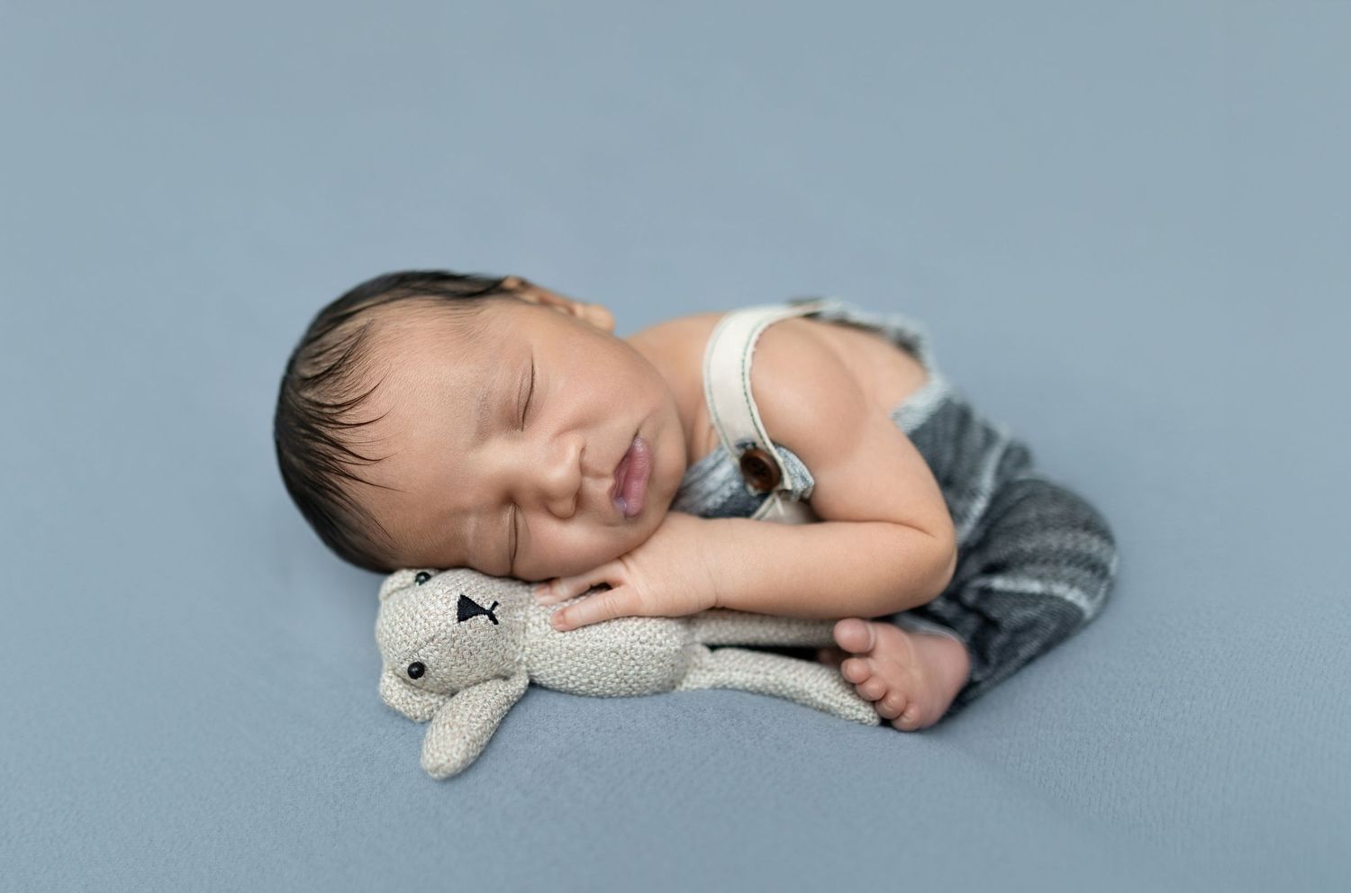 Sleeping newborn baby in gray overalls cuddling a knitted bunny toy on a soft blue background.