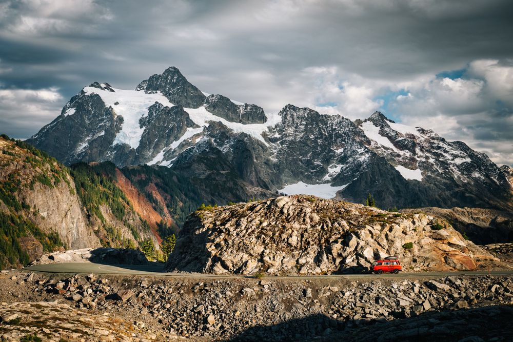 Hikers rest on rocky terrain with dramatic snow-capped mountain peaks and moody clouds in the background.