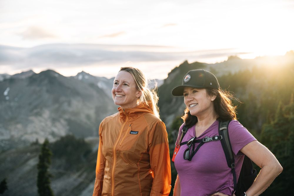 Two hikers in orange and purple jackets enjoy a beautiful sunset in the mountains with scenic mountain peaks in background.