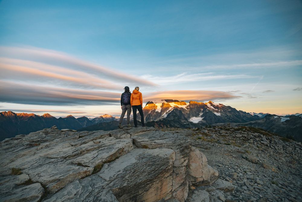 Two silhouetted figures stand on a rocky mountain summit against a dramatic sunset and mountain range backdrop.