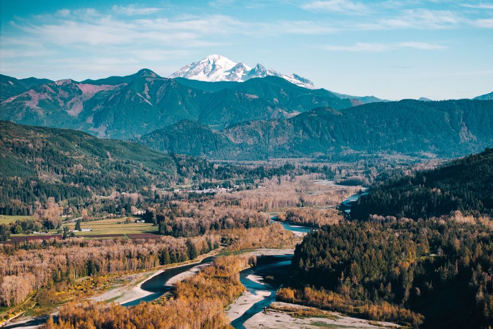 Aerial view of a winding river valley with snow-capped Mount Baker looming in the distance among evergreen mountains.