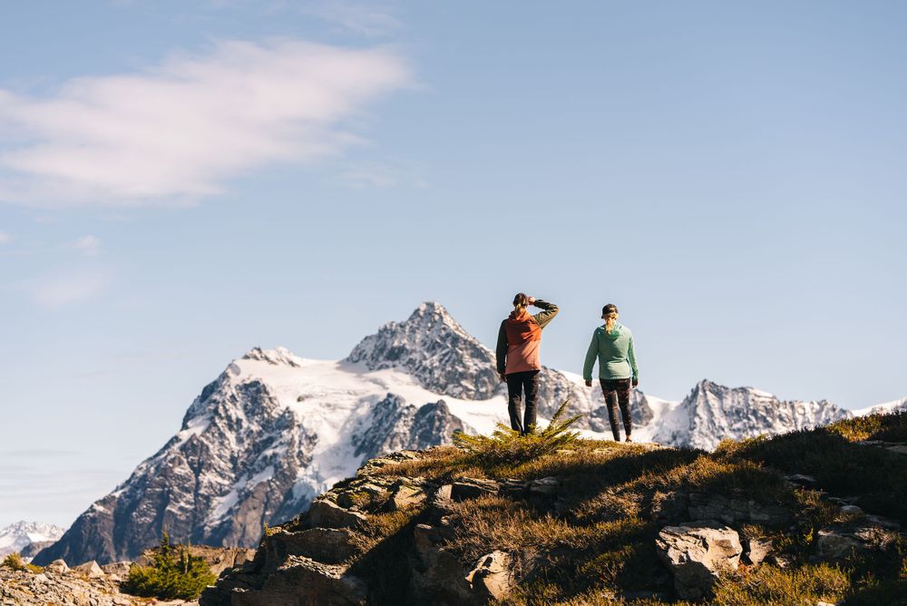 Two hikers stand on a mountain ridge overlooking snow-capped peaks against a blue sky in the Alps.