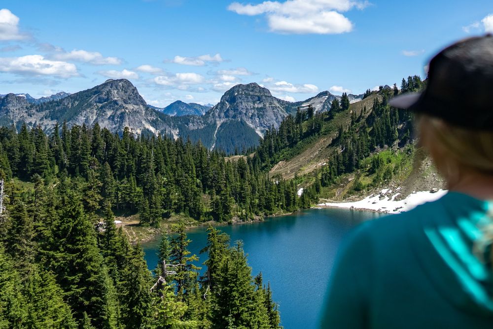 Scenic view of turquoise alpine lake surrounded by evergreen forest and rugged mountain peaks under blue sky.
