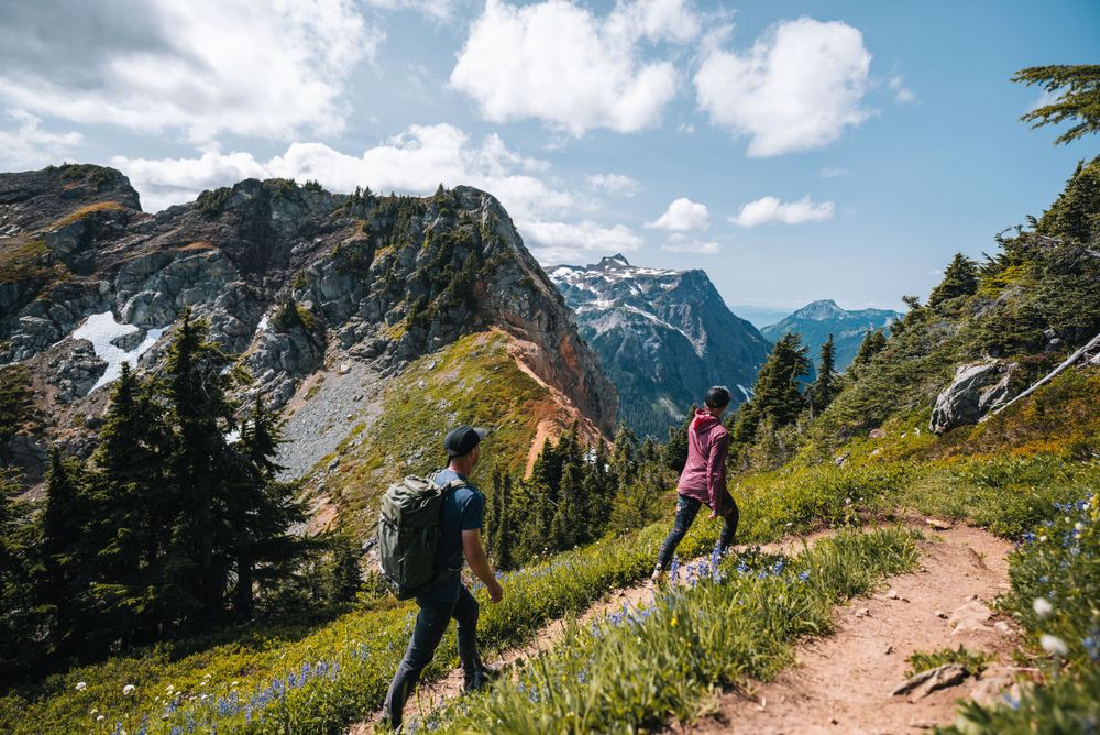 Two hikers traverse a scenic mountain trail overlooking snow-capped peaks in the North Cascades.