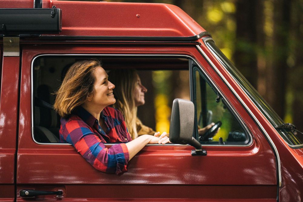 Red camper van parked in lush forest with sunlight filtering through trees.