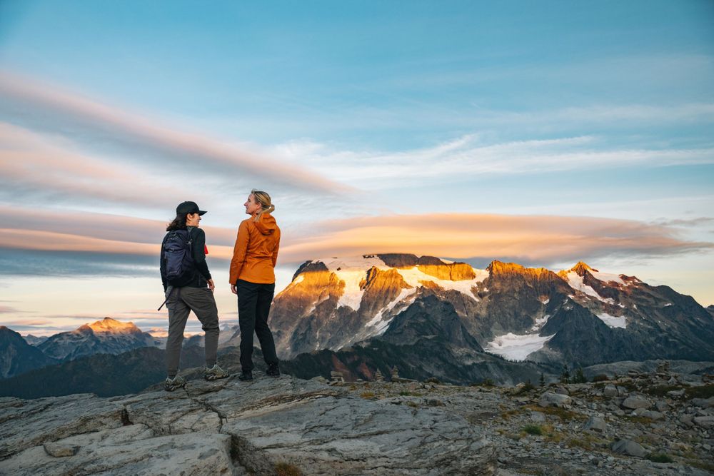 Two hikers stand on a mountain summit overlooking snow-capped peaks during a colorful sunset.