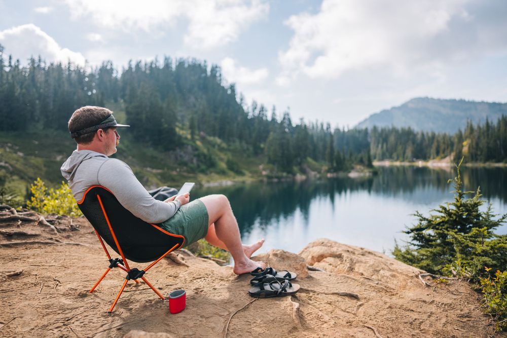 Camper relaxes in portable chair by alpine lake with mountain views and camping gear nearby.