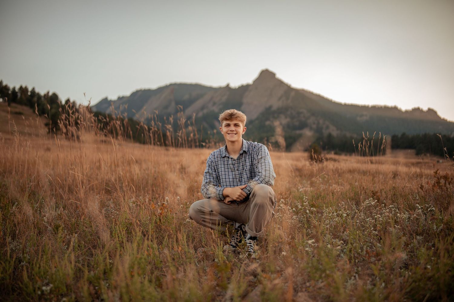 Someone sits in a field of golden grass with rugged mountain peaks and evergreen forest visible in the background.