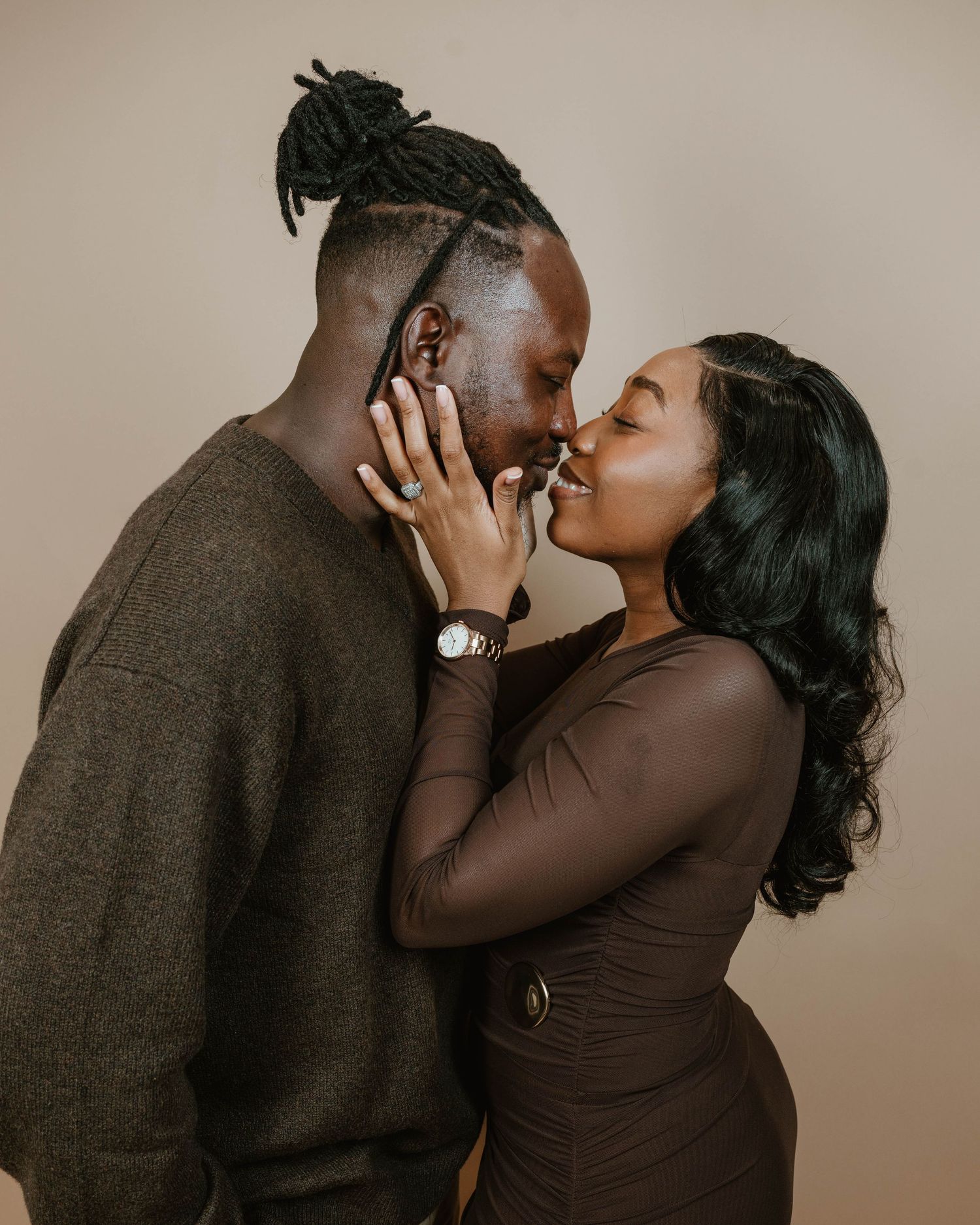 A couple sharing a romantic moment, smiling and about to kiss in a studio setting wearing coordinated brown outfits.