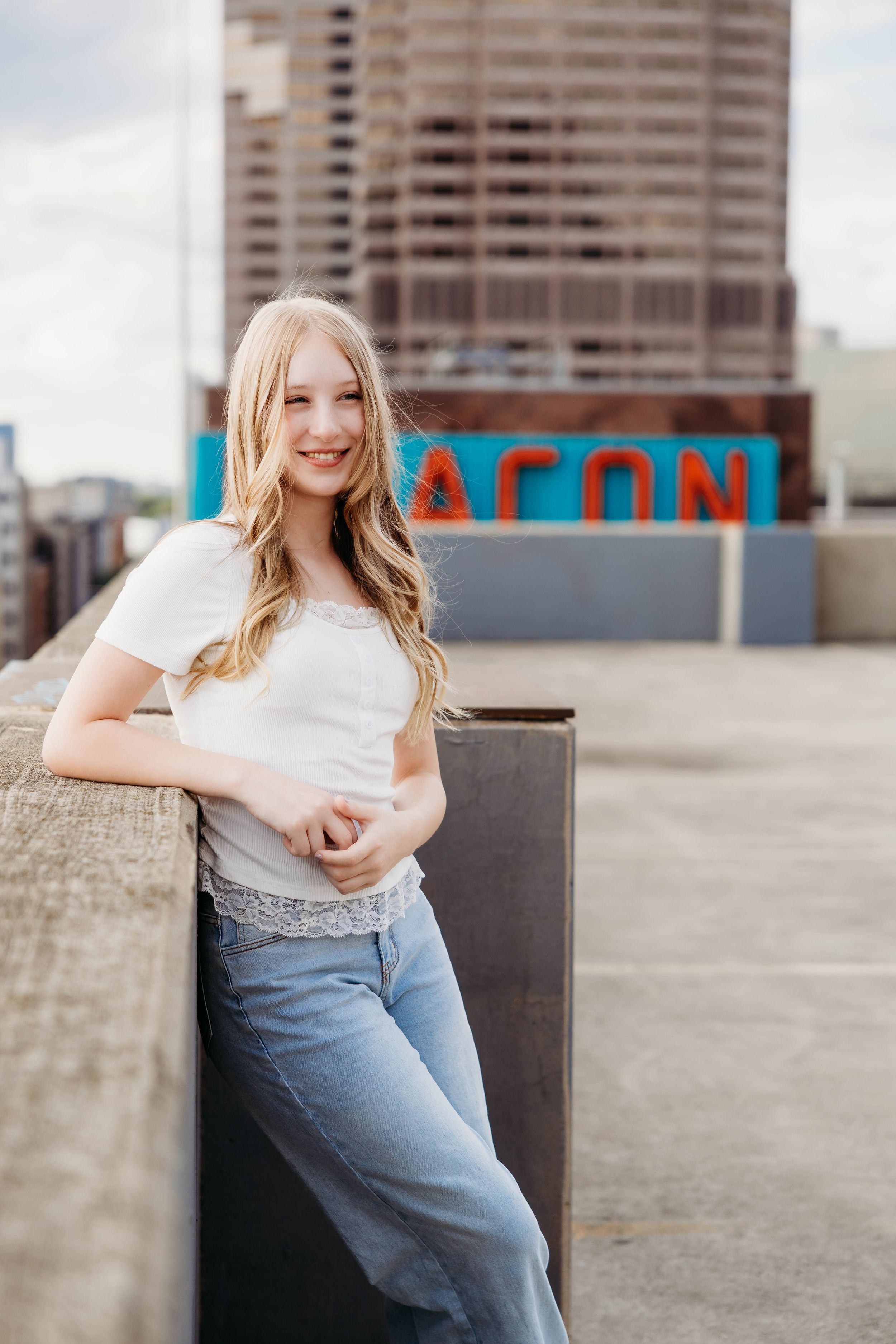 Smiling blonde teen girl in white top and jeans leaning on rooftop ledge with city buildings behind her.
