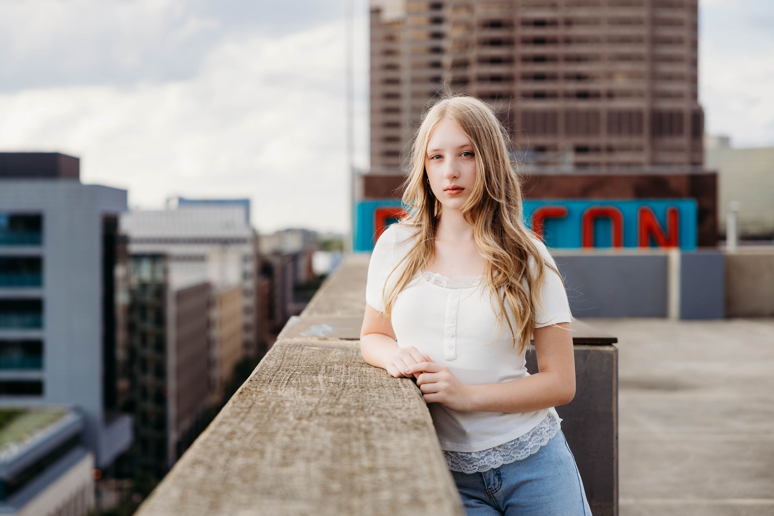 Young blonde woman in white top and jeans poses on urban rooftop with city buildings in background.